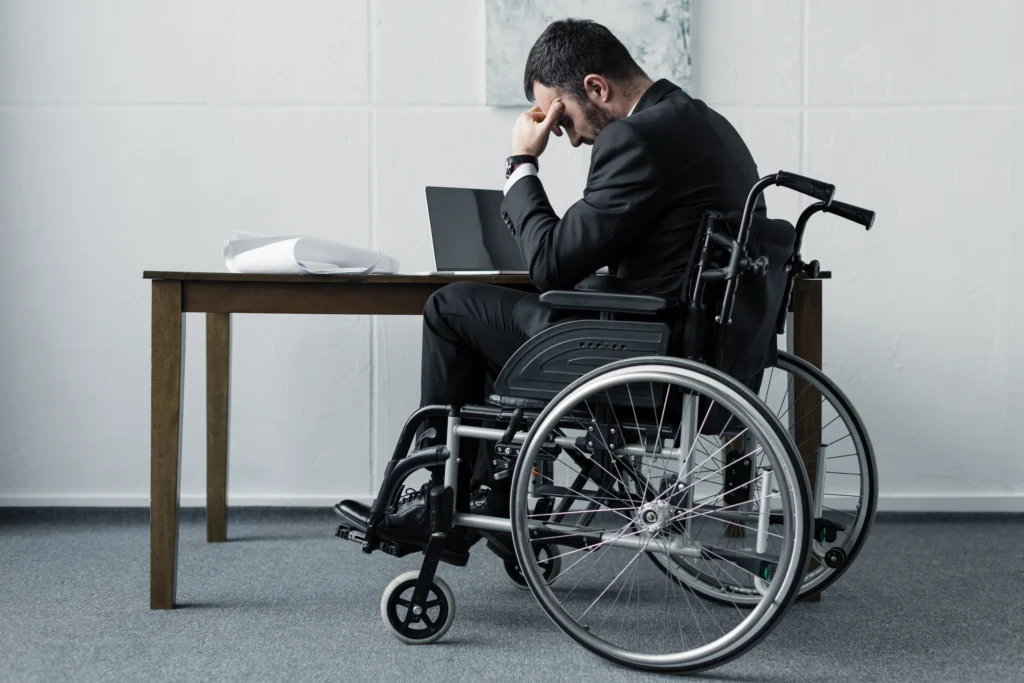 A man in a suit sits in a wheelchair at a desk with a laptop, resting his head on his hand, appearing contemplative or stressed.