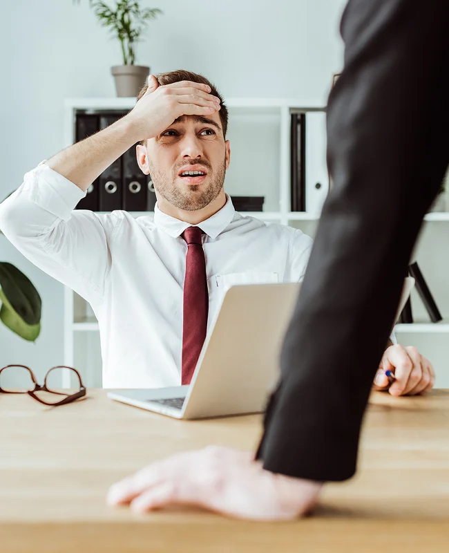 A worried man in a white shirt and red tie sits at a desk with a laptop, holding his forehead. Another person stands opposite, creating tension.