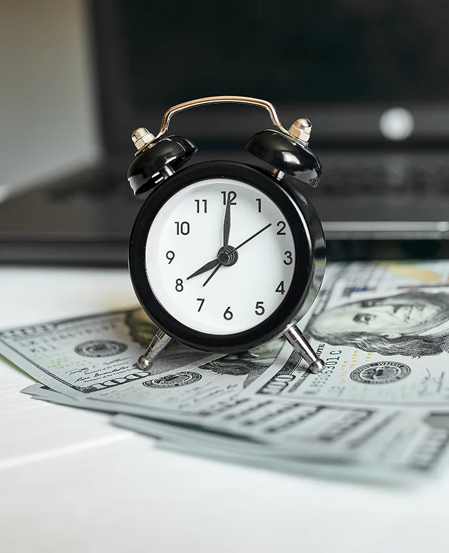 A black alarm clock sits on a stack of U.S. hundred-dollar bills with a blurred laptop in the background, symbolizing the concept of time and money.