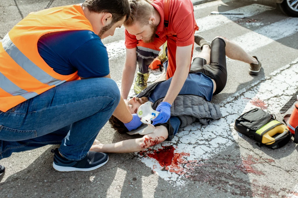 A group of people assisting a man lying on the ground in an outdoor setting. They are surrounded by vehicles, and one person is seen holding a writing implement. The scene includes a sidewalk and chalk nearby.