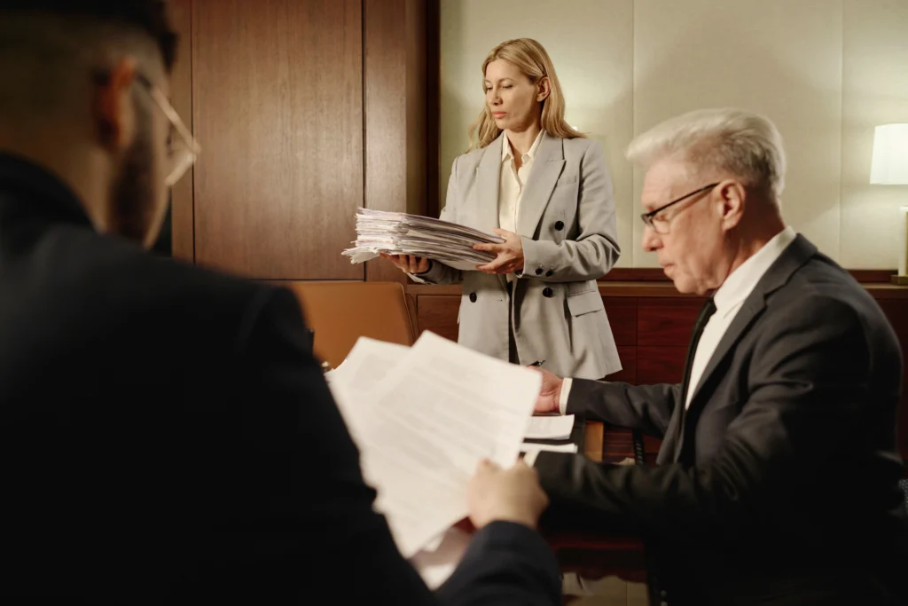 A woman with light brown hair is holding a stack of papers, standing indoors next to a wall. She is dressed in formal attire, appearing engaged in a conversation.