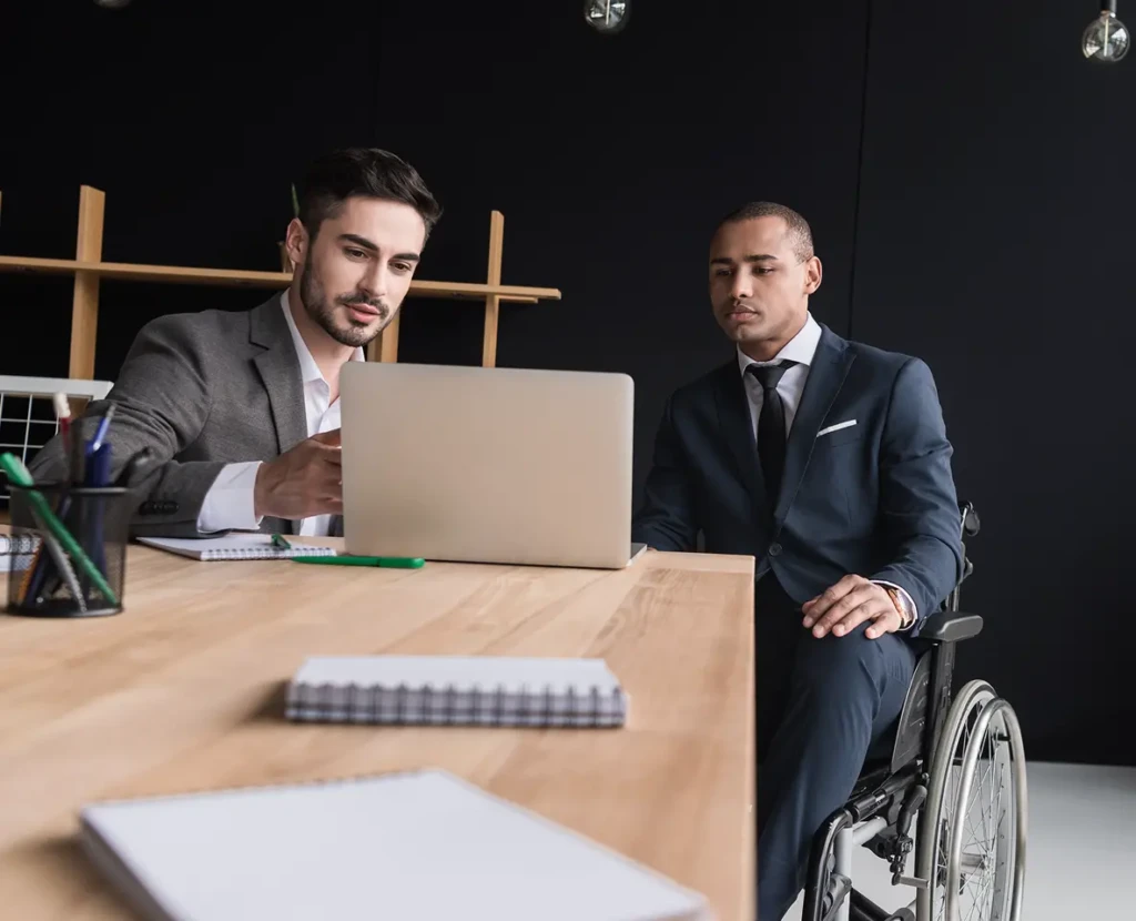 Two men in suits engage in a focused discussion at a desk with a laptop. One man is in a wheelchair, conveying inclusivity in a modern office.